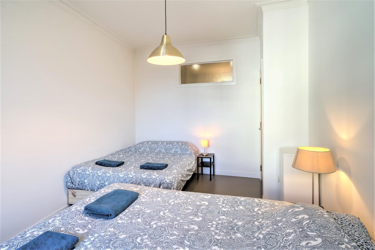 A room with two beds, a hanging light fixture, a small table with a lamp, and a wall-mounted light.