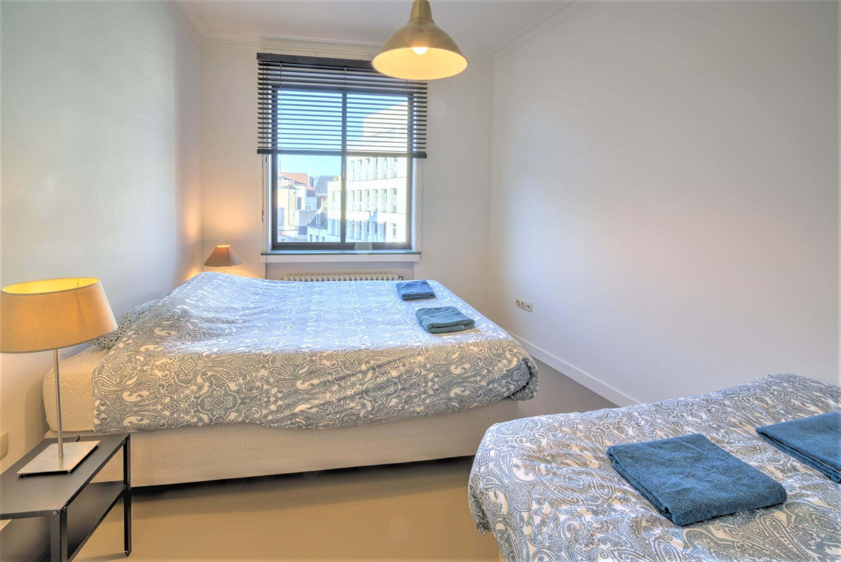Un dormitorio con cama, mesita de noche, lámpara y ventana con persianas.