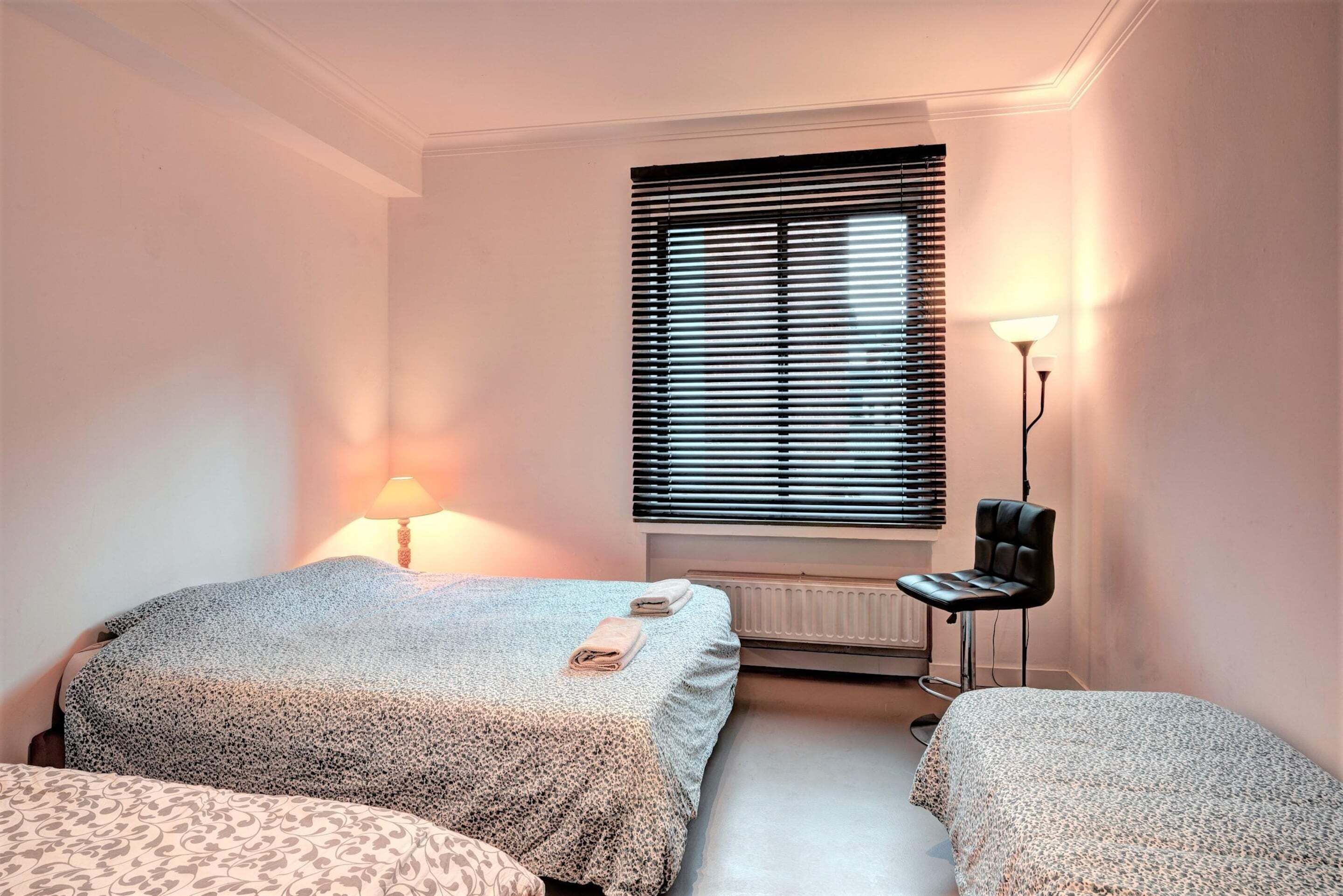 Un dormitorio con dos camas, un radiador, una ventana con persianas, un escritorio con silla y una lámpara.