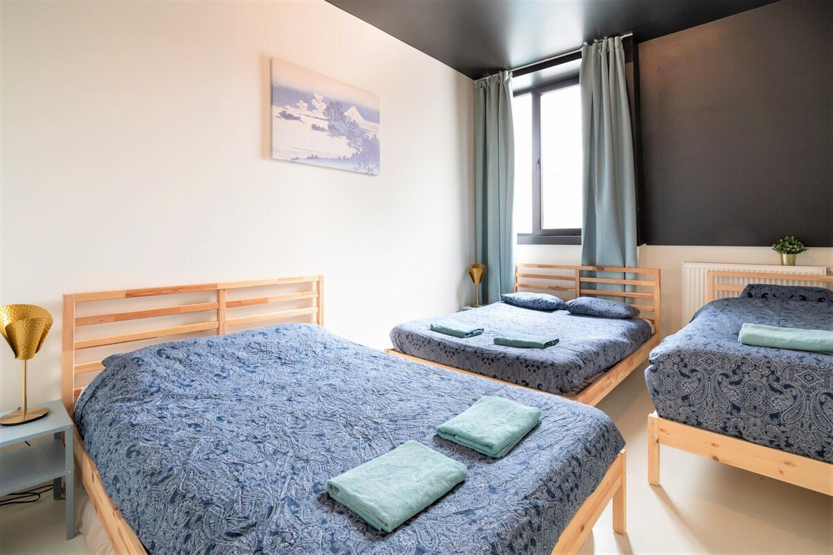A room with three beds, a window with curtains, a painting on the wall, and a small plant on a table.
