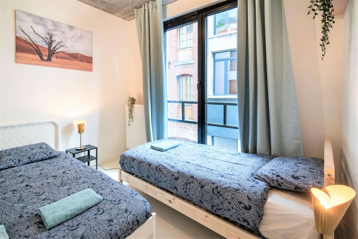A bedroom with two beds, a window with curtains, a bedside lamp, and a wall-mounted plant.