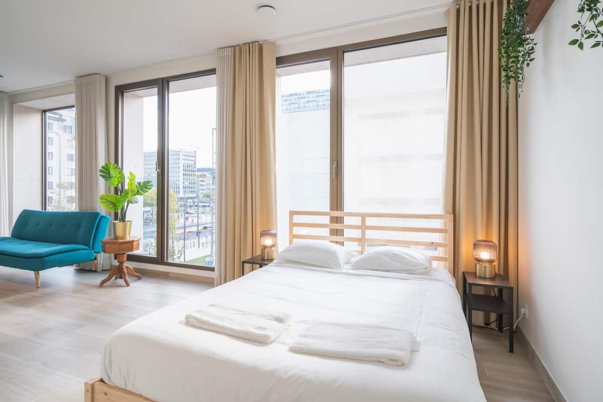 Une chambre d’hôtel avec un grand lit, une chaise bleue, une petite table avec une plante, et une vue sur les bâtiments à travers de grandes fenêtres.