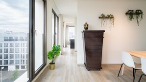 Una habitación de hotel moderna con ventanas amplias, un aparador de madera, una mesa de comedor con sillas y plantas colgantes.