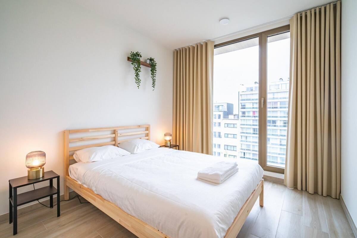 Une chambre à coucher avec un lit en bois, des lampes de chevet, une petite table, une fenêtre avec des rideaux et une vue sur les bâtiments à l’extérieur.