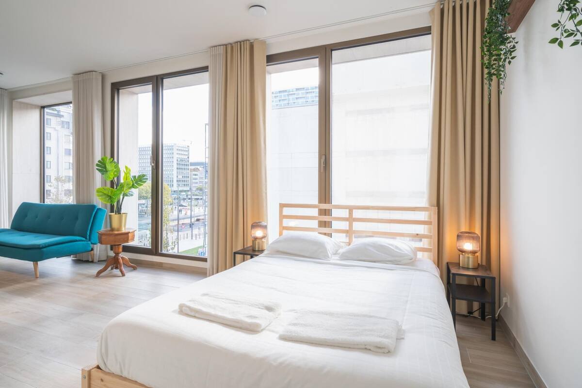 Une chambre d’hôtel avec un grand lit, une chaise bleue, une plante en pot et une vue sur la ville.