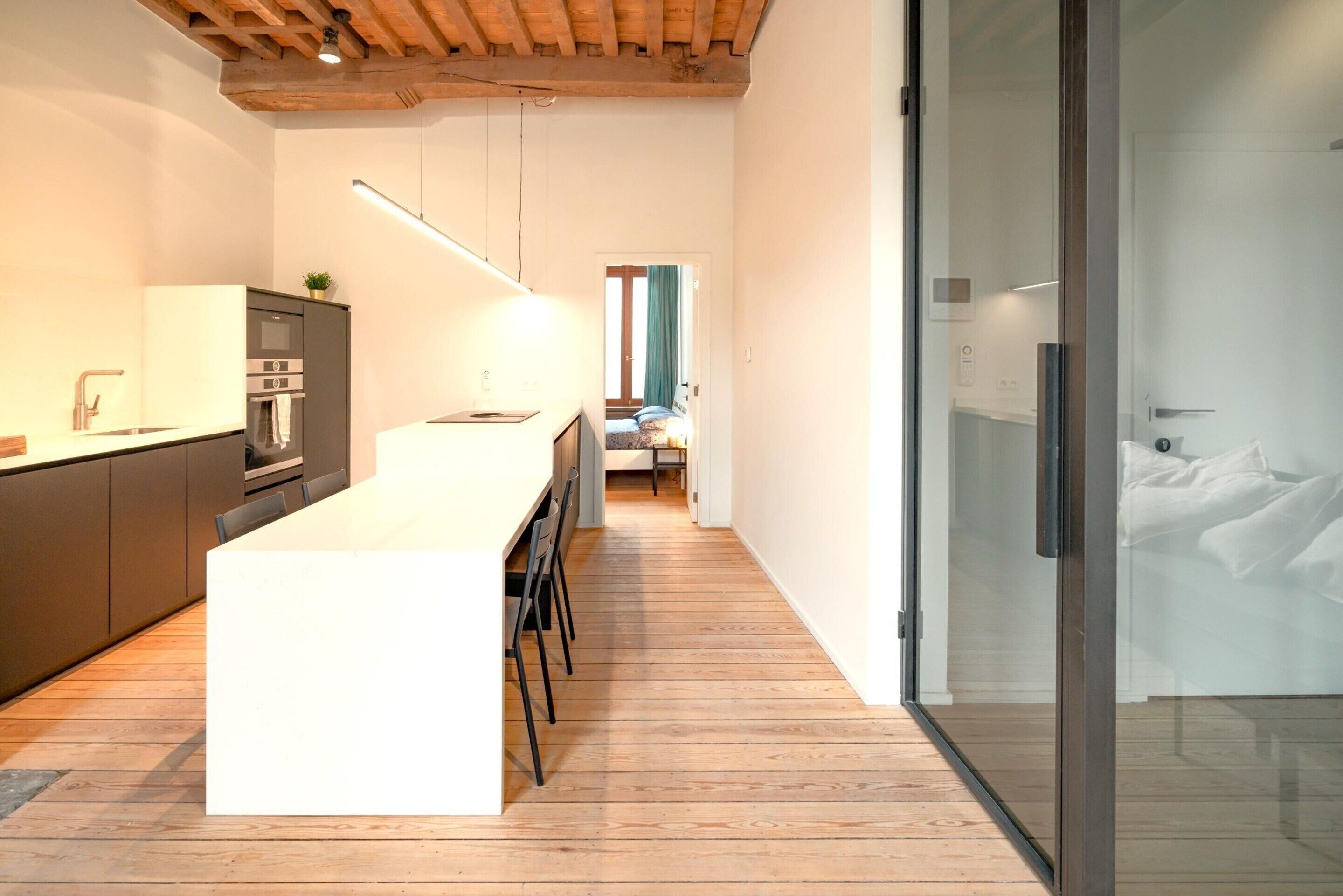 A modern kitchen with a central island, wooden flooring, and a glass door leading to a bedroom.