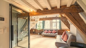 Un cuarto tipo loft con techo de madera, una puerta corrediza de vidrio que da a un balcón, un sofá con cojines y una mesa de comedor.