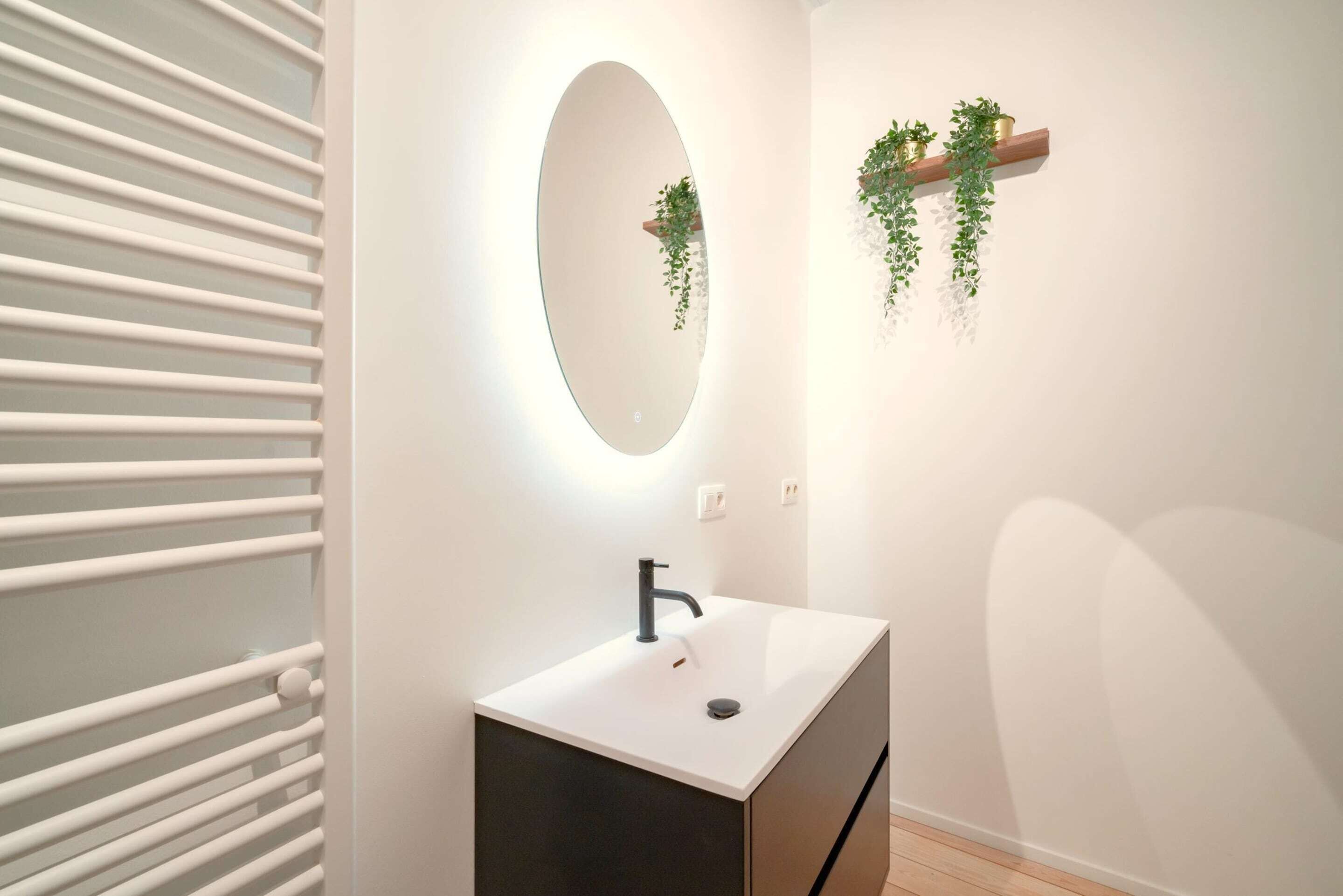 Un baño con un lavamanos blanco, un espejo, un estante de madera con plantas y un radiador blanco.
