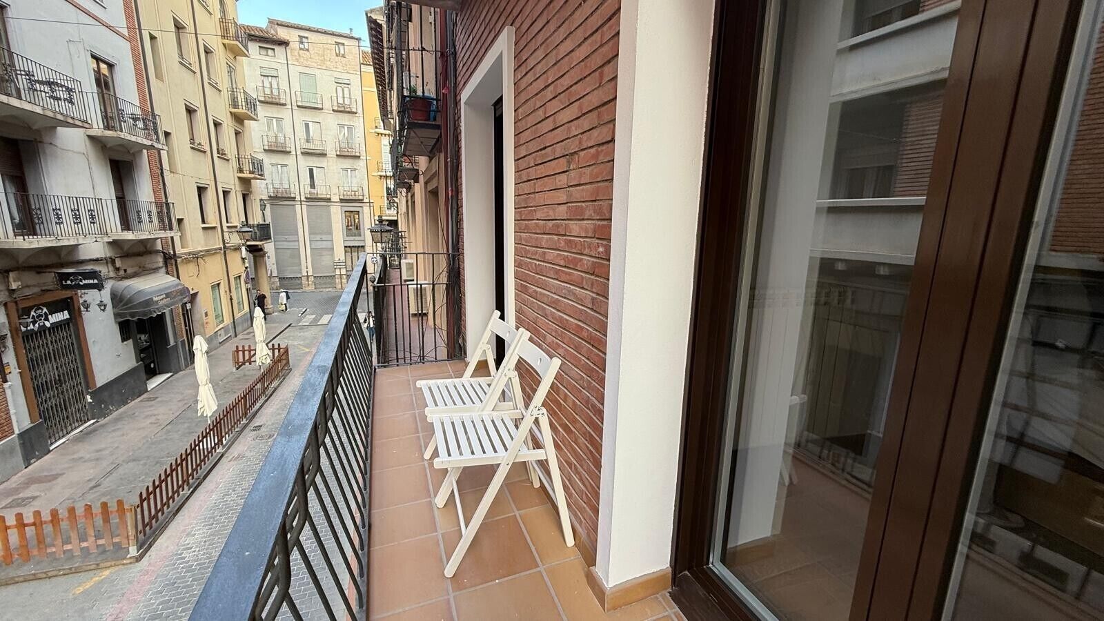 Un balcón estrecho con suelo de baldosas, pared de ladrillo y puerta de cristal que da a una habitación.