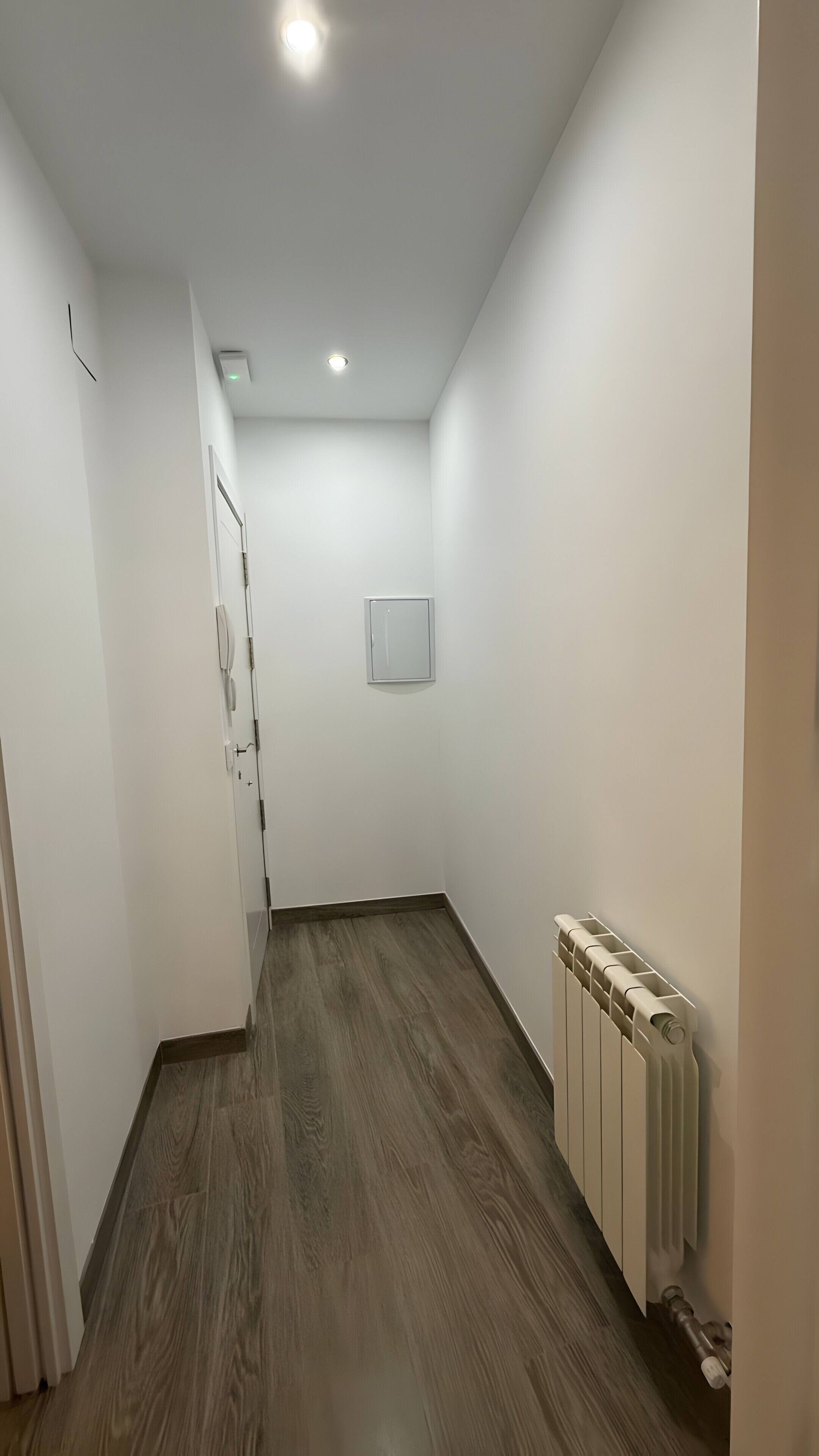 Un pasillo con suelo de madera, paredes blancas, un radiador y una puerta con picaporte.