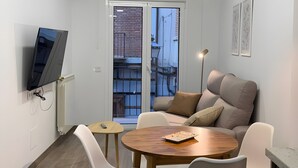 A modern dining area with a round wooden table, white chairs, a black pendant light, and a flat-screen TV mounted on the wall.