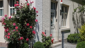 Um edifício com fachada de pedra, um pequeno jardim com flores cor-de-rosa e um caminho de paralelepípedos.