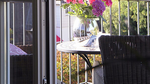 Un balcón con una silla de mimbre, una mesita y un jarrón de flores.