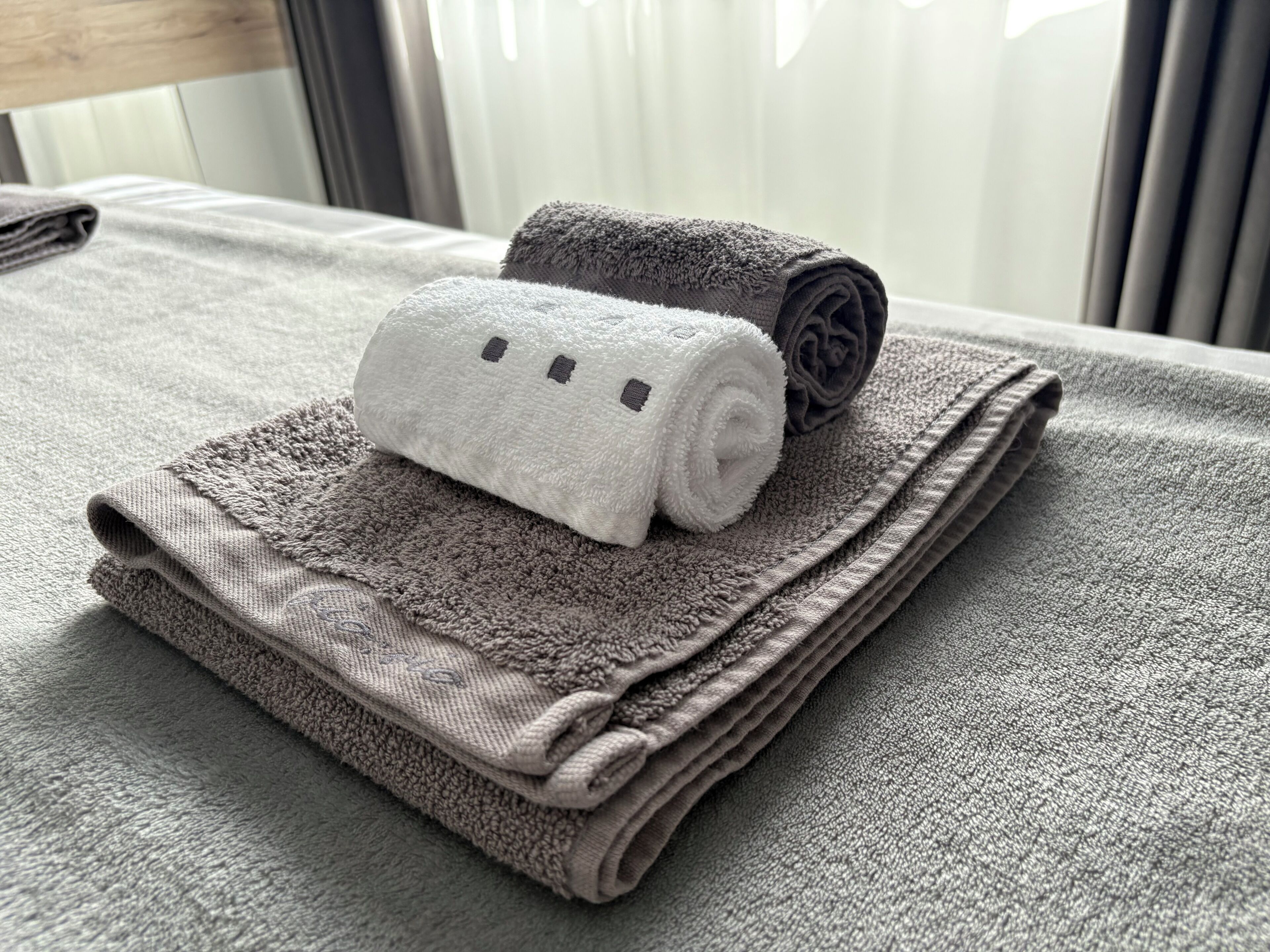 A neatly folded set of towels on a bed with a headboard and curtains in the background.