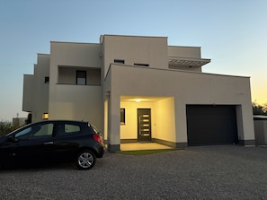 A modern two-story building with a car parked in front, a garage, and a well-lit entrance. - Cozy family apartment in quiet surroundings close to the sea (Kostrena)