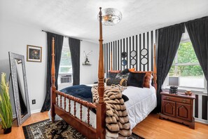 A four-poster bed with a fur throw, a wooden nightstand, a mirror, and a window with curtains.