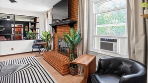 Ein Wohnzimmer mit Kamin aus Backstein, einem schwarzen Ledersessel, einem Holztisch und einem großen Fenster mit Vorhängen.