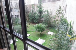 Blick durch ein Fenster auf einen Innenhof mit einem kleinen Baum, einem Busch und einem Gebäude im Hintergrund.