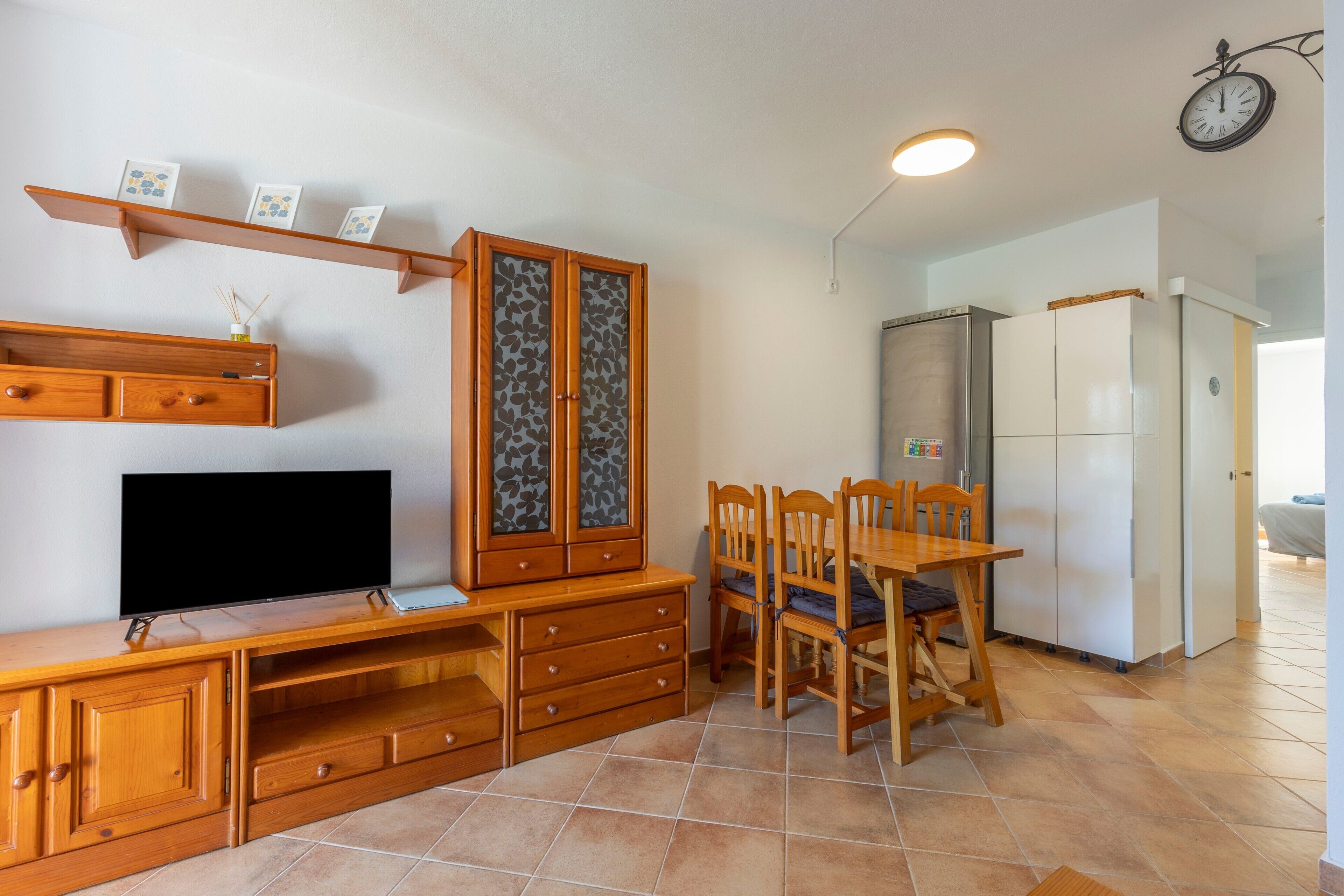 A compact kitchen with a dining table, chairs, a TV, wooden cabinets, and a refrigerator.