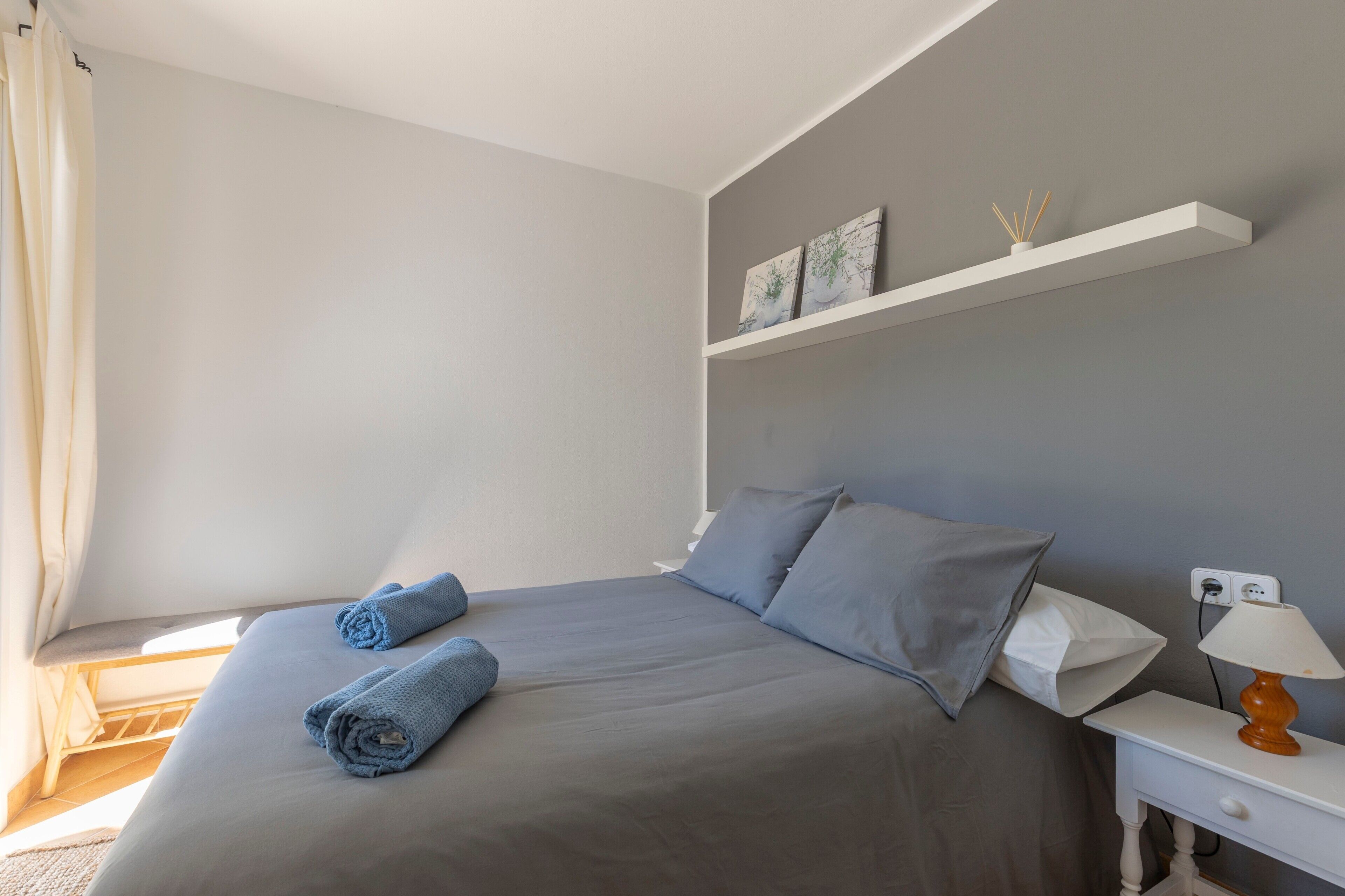 Une chambre à coucher avec un lit, des oreillers, une table de chevet avec une lampe et une étagère avec des œuvres d’art.