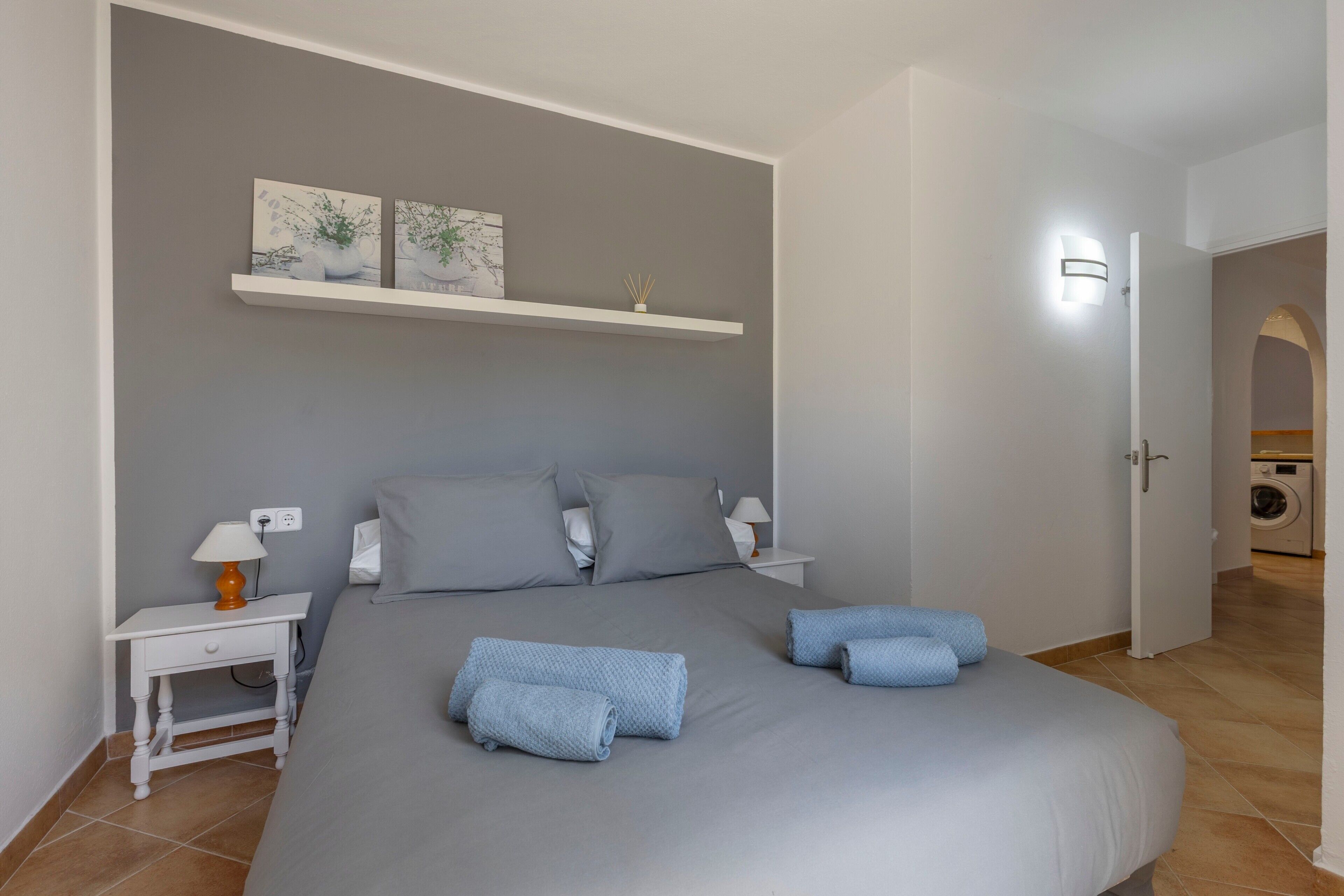 Ein Schlafzimmer mit einem Bett, zwei Nachttischen, einer Wandablage mit Dekoration und einer Tür zu einem Waschbereich.