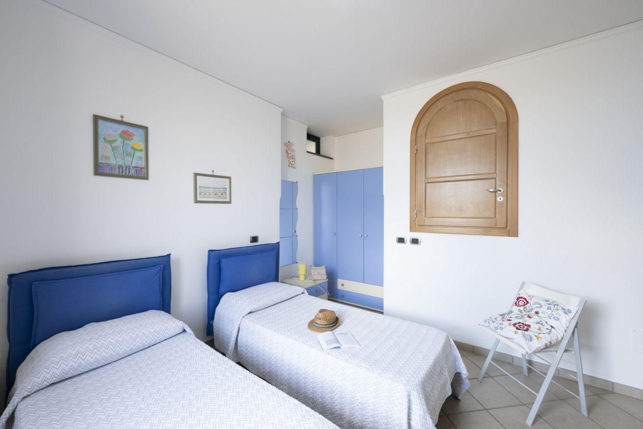 A bedroom with two beds, a wooden door, a small table with a chair, and a wall with framed pictures.
