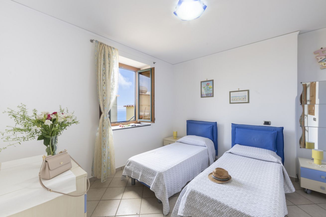 A bedroom with two beds, a window with curtains, a bedside table, a chair, and a vase with flowers.