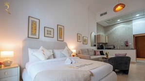 Quarto de hotel com cama, mesas de cabeceira, sofá, poltrona, área de cozinha e obras de arte emolduradas na parede.
