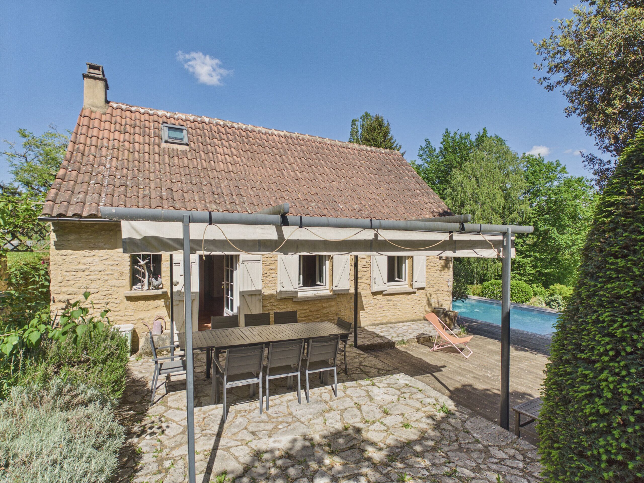 A stone house with a tiled roof, a covered patio with a table and chairs, and a swimming pool.