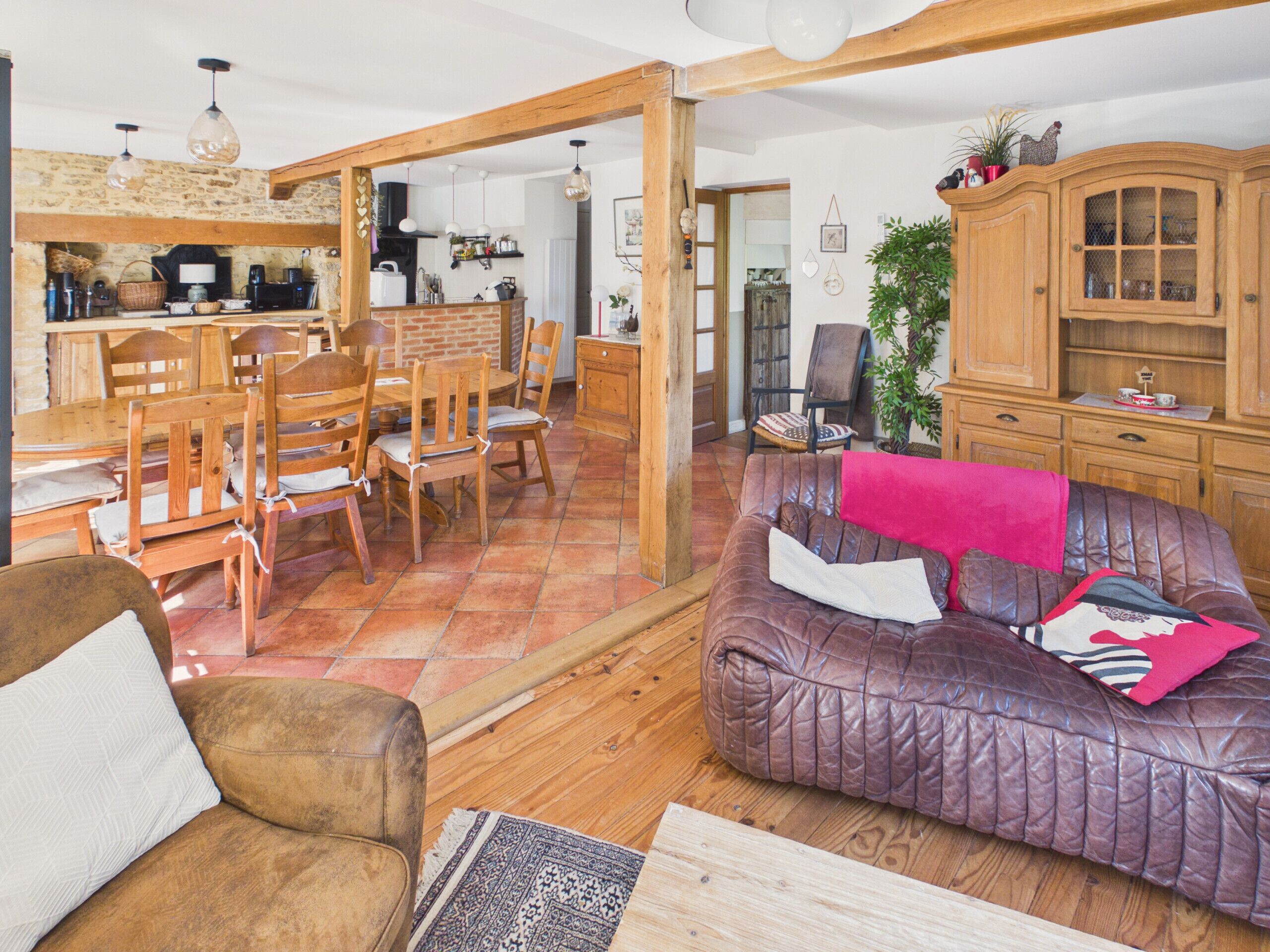 Una sala de estar acogedora con un sofá de cuero marrón, una mesa de comedor de madera y sillas, una zona de cocina con pared de ladrillo y un mueble de madera con puertas de vidrio.