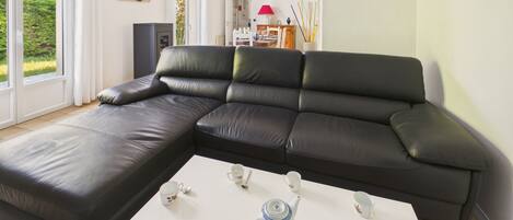 A modern living room with a black leather sectional sofa, a white coffee table with tea set, and a view of the outdoors through large windows.