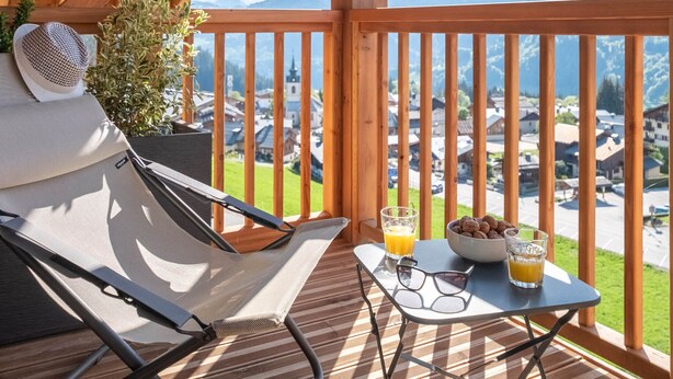 Un terrazzo in legno con un'amaca, un tavolo con bicchieri e bevande, e vista su un villaggio e montagne.