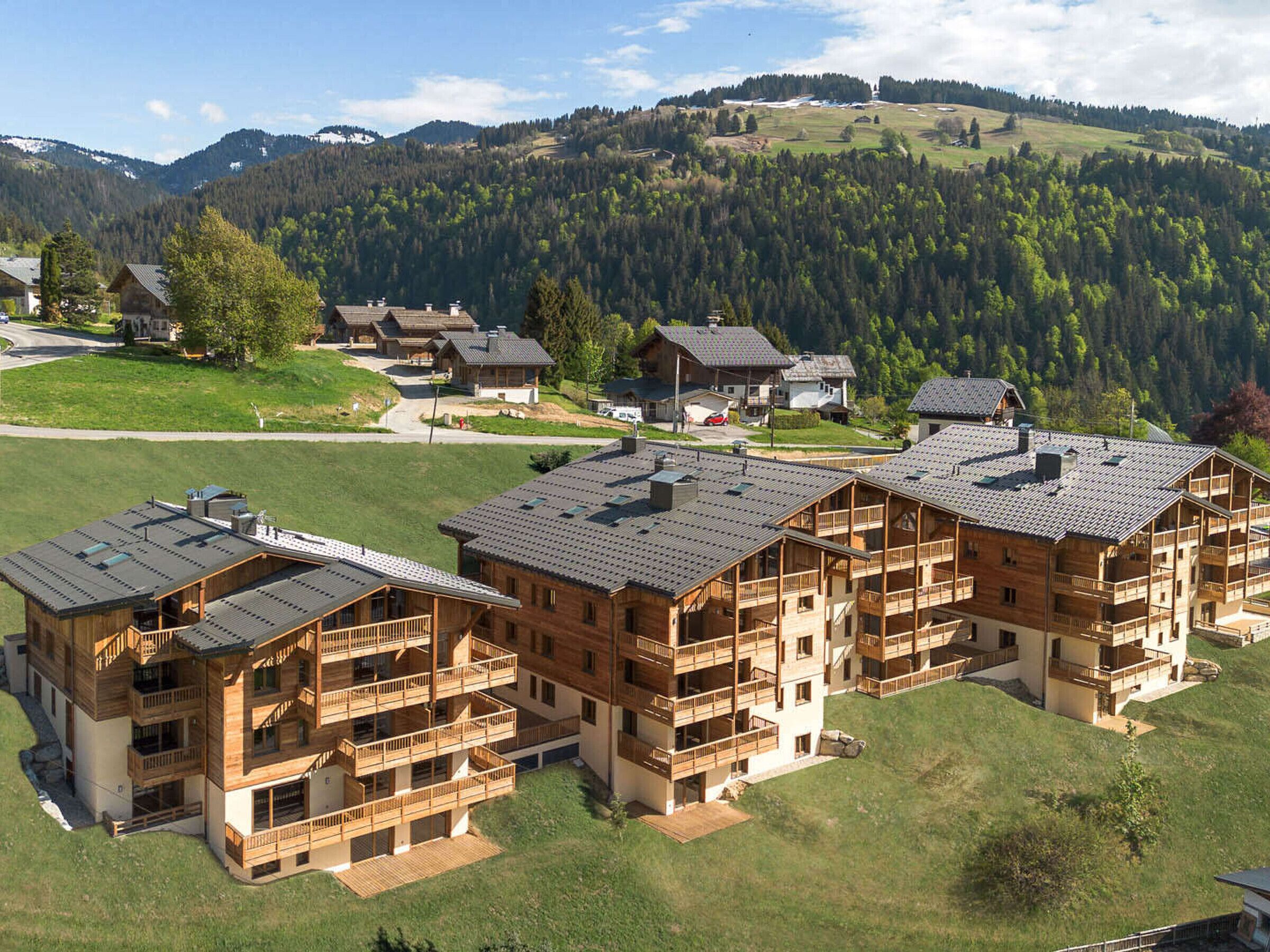 A cluster of wooden multi-story buildings with balconies, set in a hilly area with a forested backdrop.