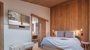 Una camera da letto con soffitto in legno, un letto con testiera bianca, una panca e una TV a parete che mostra una scena di bosco innevato.
