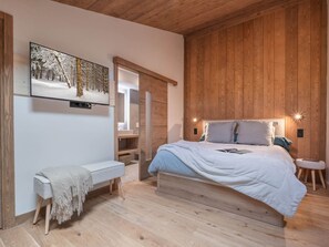 Ein Schlafzimmer mit Holzbalkendecke, einem Bett mit weißem Kopfteil, einer Bank und einem an der Wand befestigten Fernseher, der eine verschneite Waldszene zeigt.