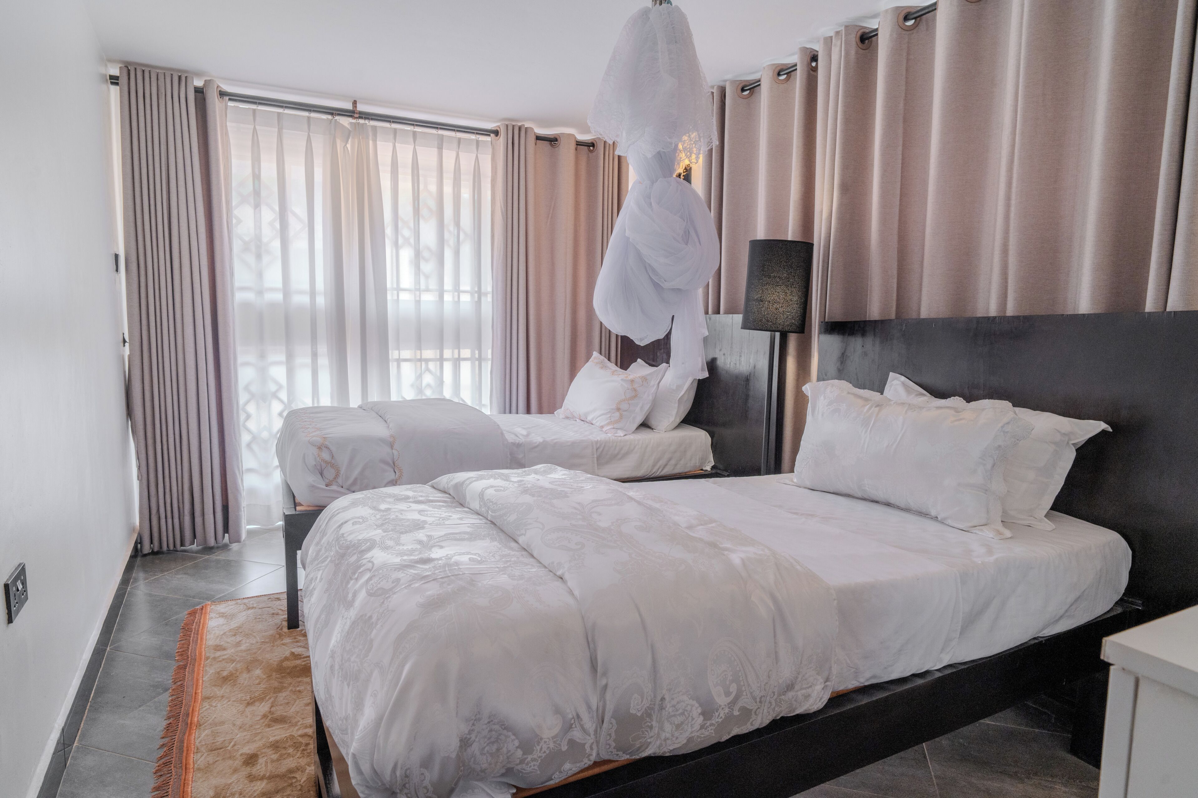 A hotel room with two beds, a large window with curtains, a nightstand, and a lamp.