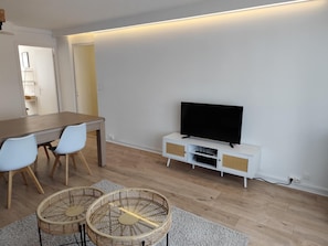 Una sala de estar moderna con una mesa de comedor de madera, dos sillas blancas, un televisor de pantalla plana sobre un mueble blanco y dos cestos de mimbre sobre una alfombra gris.
