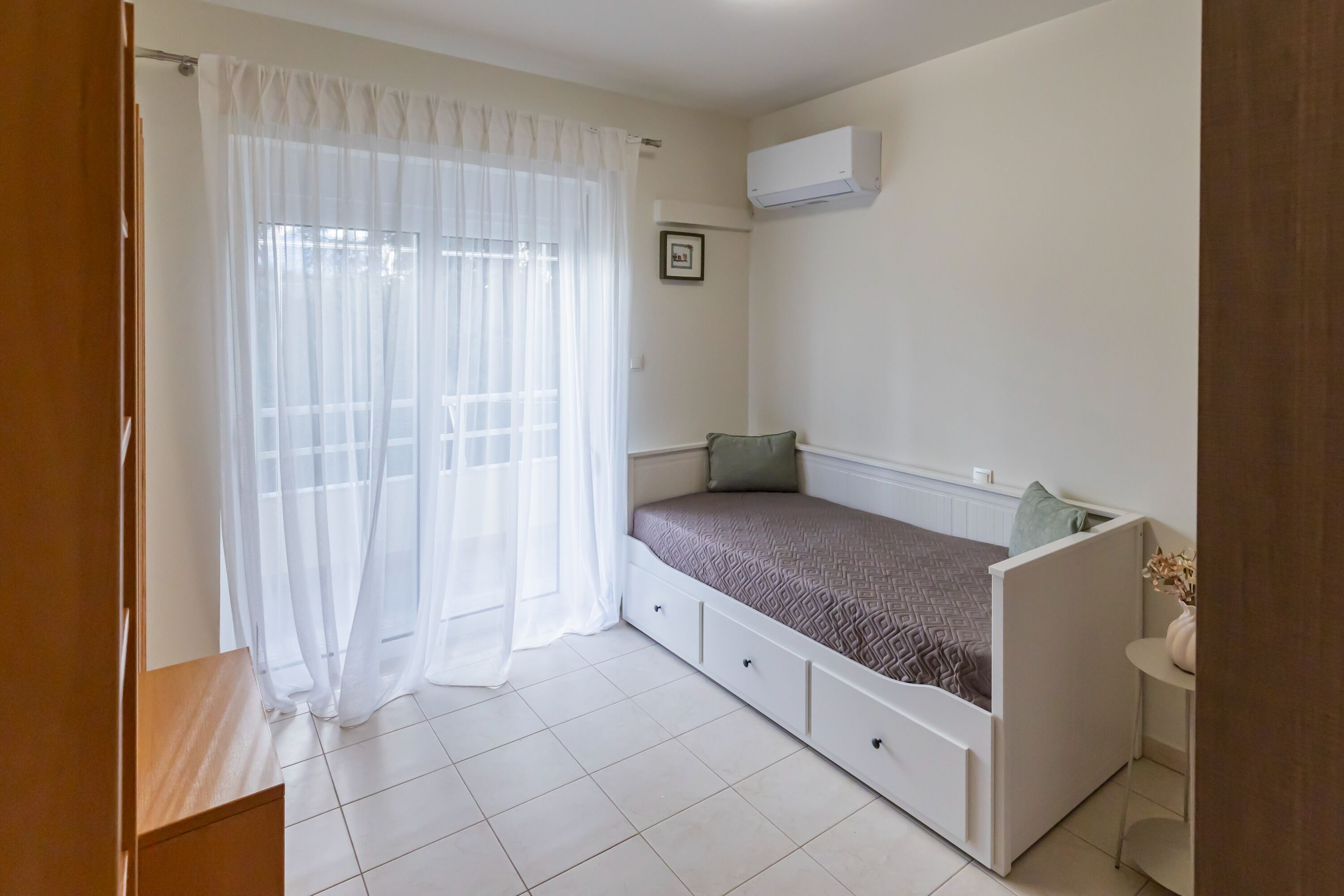 A bedroom with a bed, a small table, a wall-mounted air conditioner, and a window with sheer curtains.