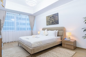 A modern hotel room with a large bed, bedside tables, a nightstand, a ceiling light, a window with curtains, and a wall-mounted artwork.