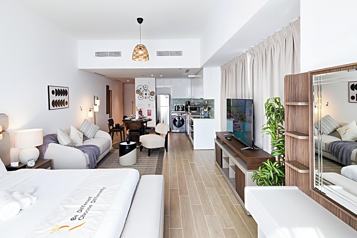 A modern hotel room with a bed, a seating area, a TV, and a kitchen area.