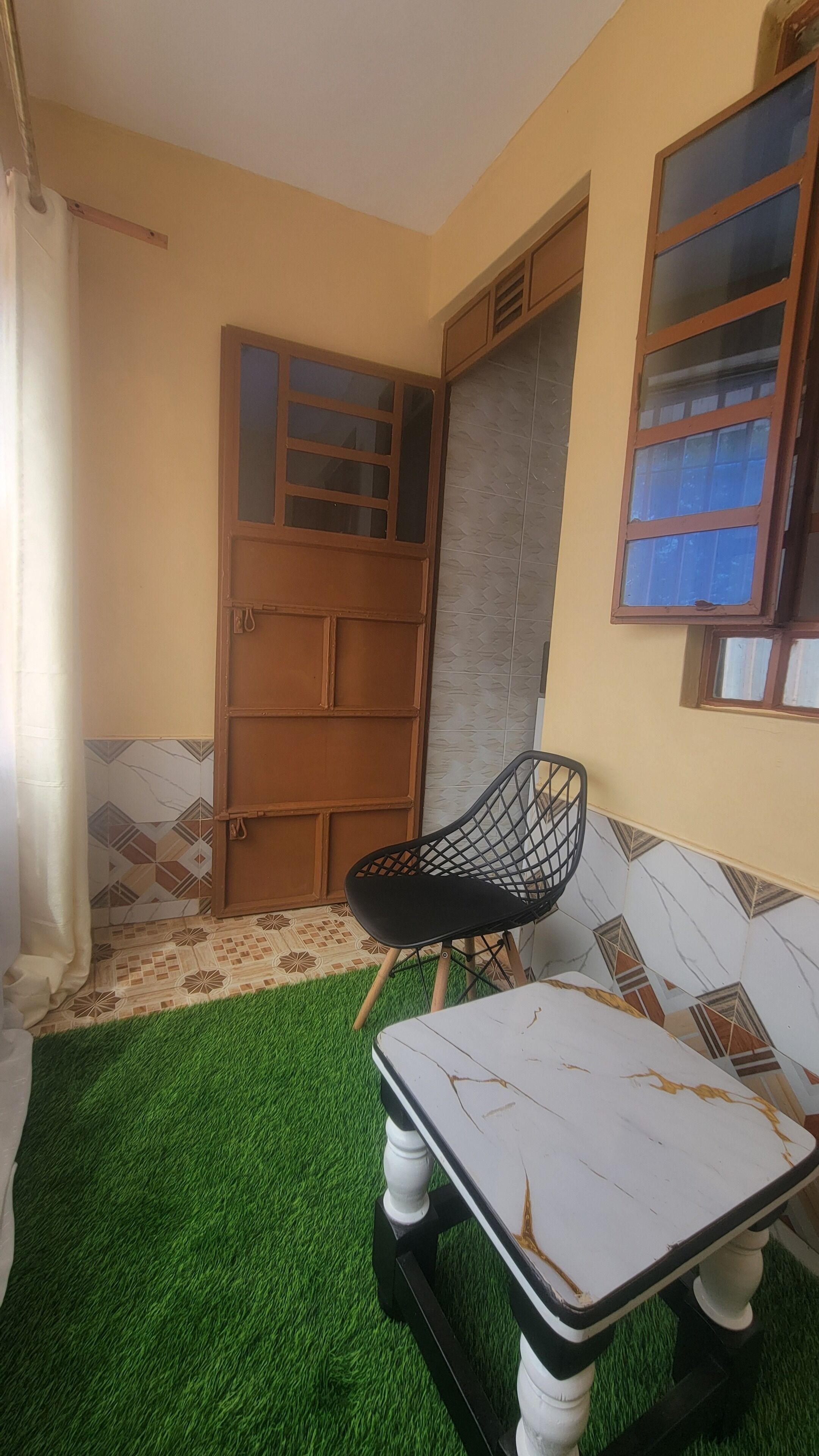 A room with a green artificial turf floor, a white marble table, a black chair, a wooden door, and a window with a view.