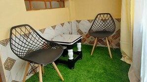 A small room with a green artificial turf floor, two black chairs with wooden legs, a small table, and a window with wooden frames. - Tranquil studio apartment in welcoming Thika neighborhood. (Thika)