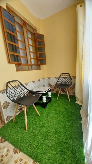 Una habitación pequeña con suelo de césped artificial verde, dos sillas negras con patas de madera, una mesita y un ventanal con marcos de madera.