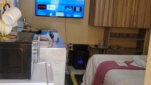 A compact hotel room with a bed, a desk with a computer, a TV mounted on the wall, and a small kitchenette area. - Tranquil studio apartment in welcoming Thika neighborhood. (Thika)