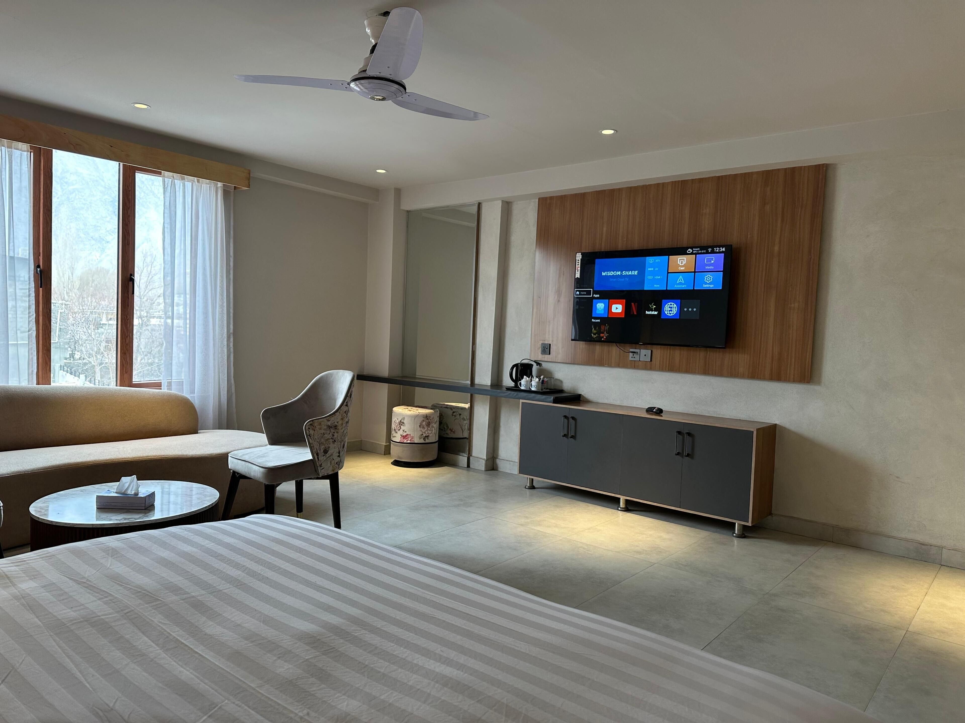 A hotel room with a bed, a sofa, a chair, a small table, a television mounted on the wall, and a ceiling fan.