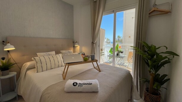 Ein Schlafzimmer mit einem Bett, einem Laptop, einem Kopfkissen mit einem Logo, einem Nachttisch, einer Pflanze und einem Balkon mit Gartenmöbeln.