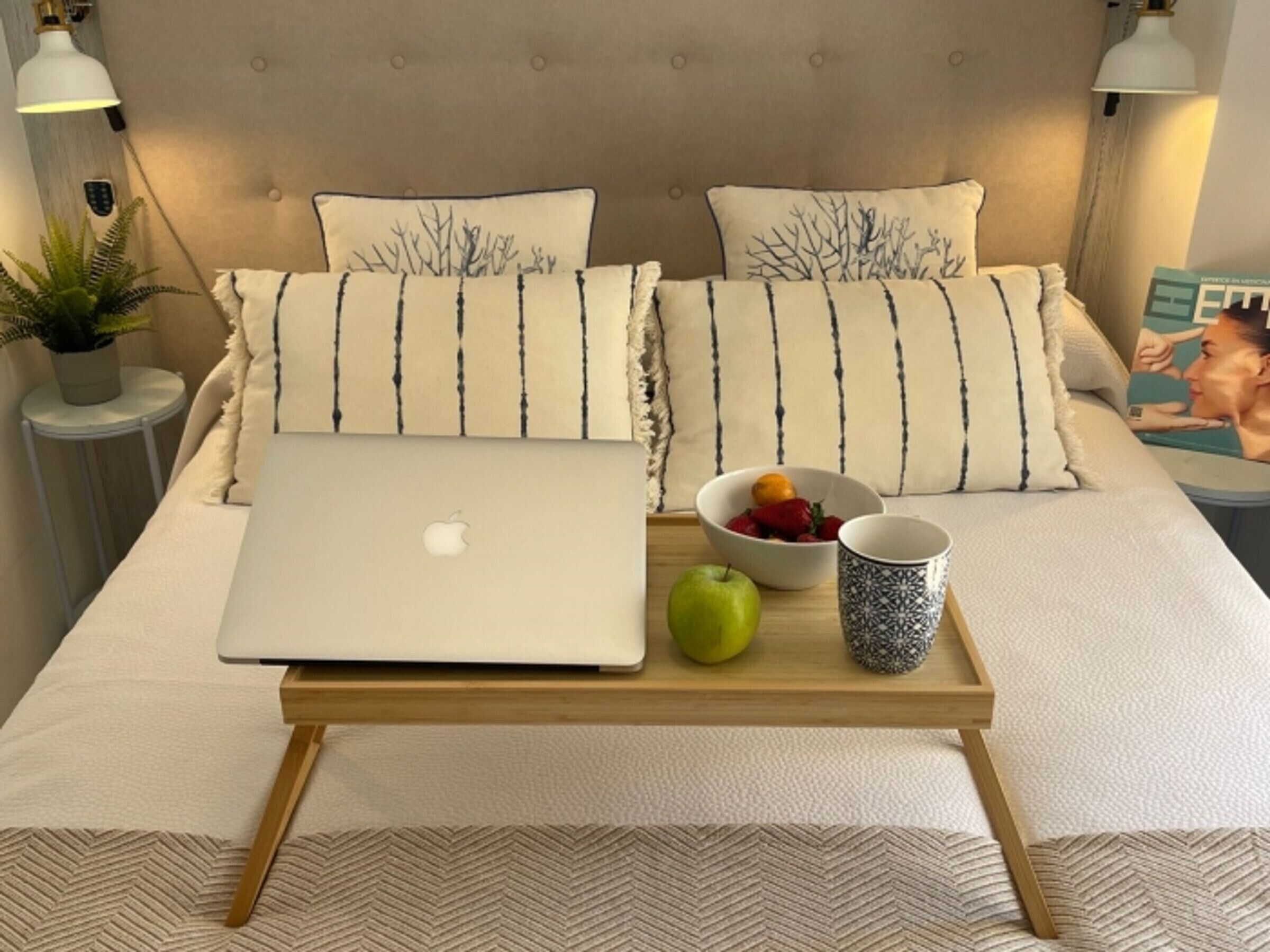 A neatly made bed with a laptop, a bowl of fruit, and a mug on a tray.