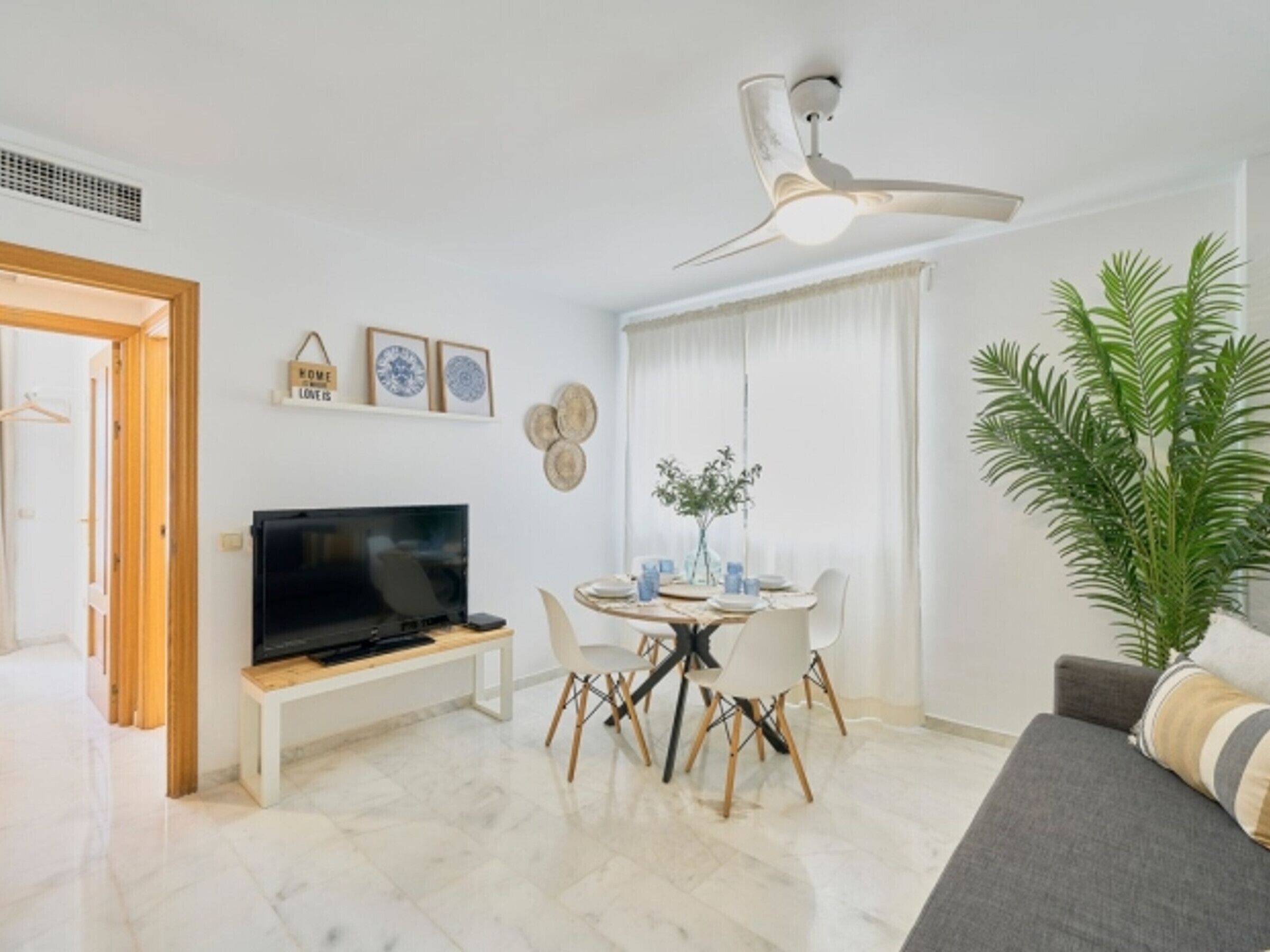 Una sala moderna con un televisor de pantalla plana, una mesa de comedor con sillas, un sofá, un ventilador de techo y una planta grande en maceta.