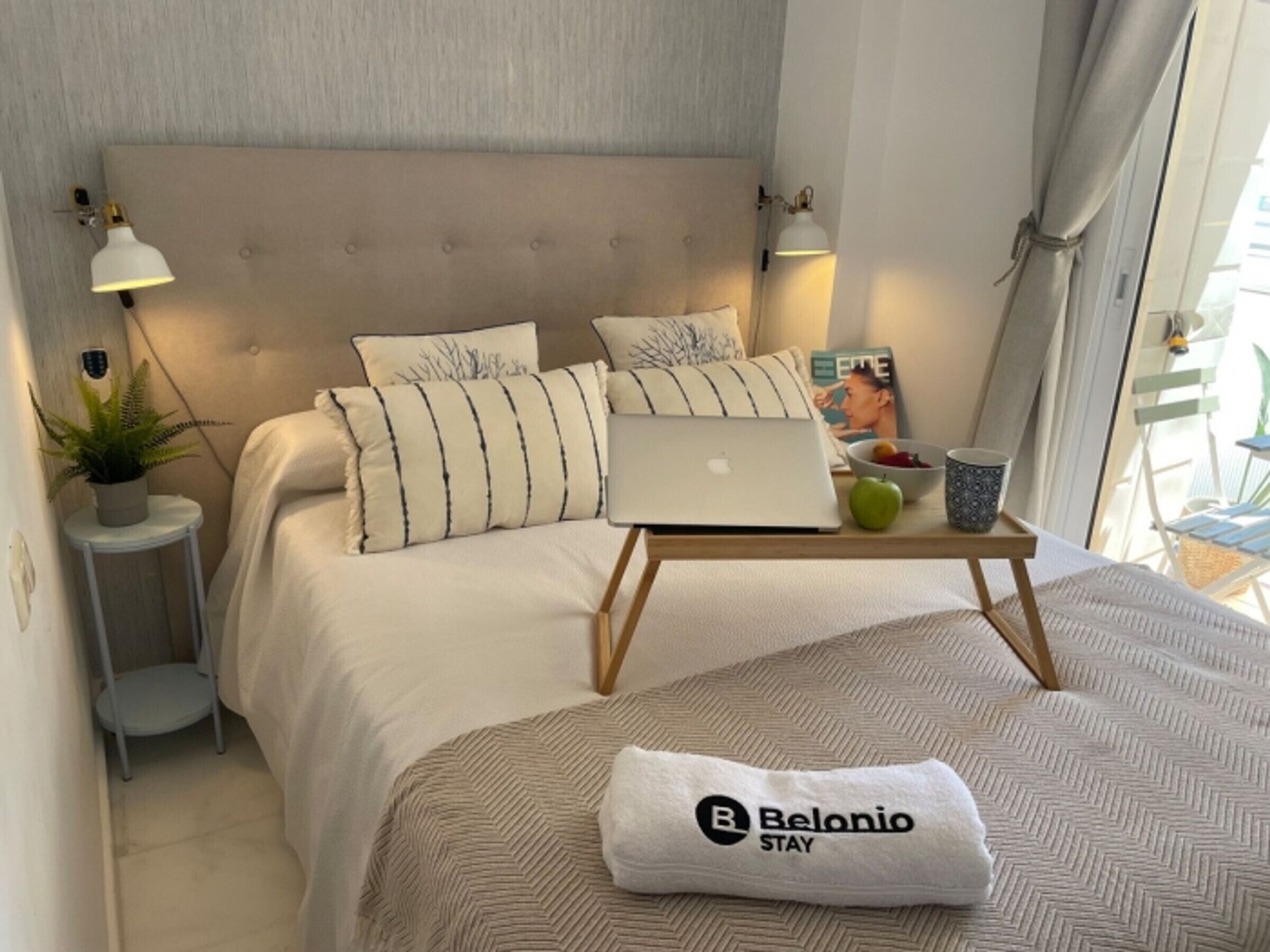 A neatly arranged bedroom with a bed, a laptop on a bedside table, a towel, and a magazine.