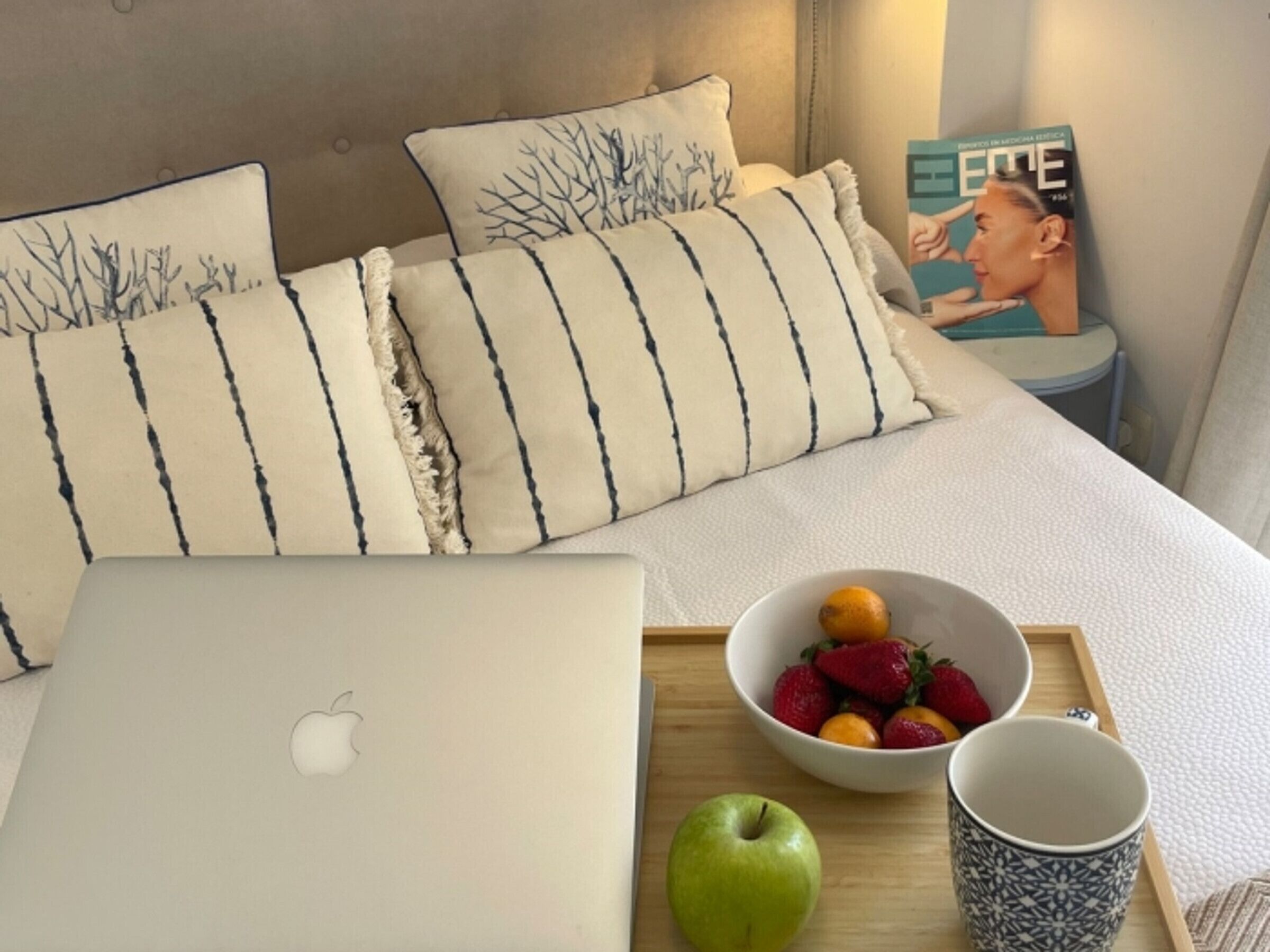 Ein ordentlich bezogenes Bett mit gestreiften Kissen, ein Laptop, eine Obstschale und eine Tasse auf einem Tablett.